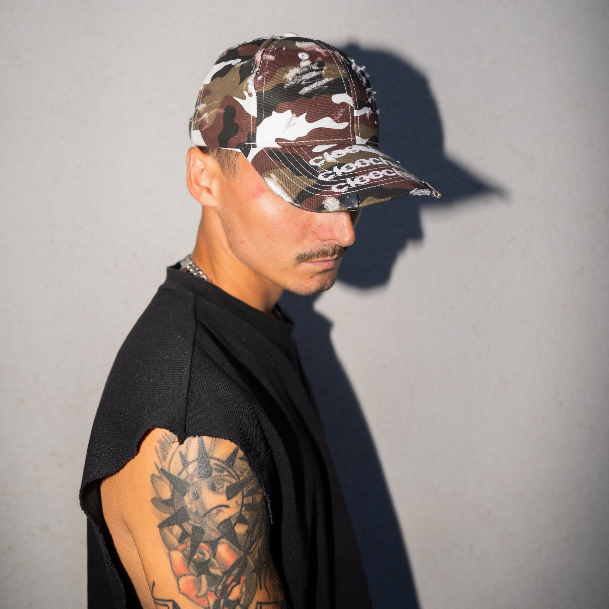 Person wearing a camouflage cap with text and tattoos on arms against a gray background