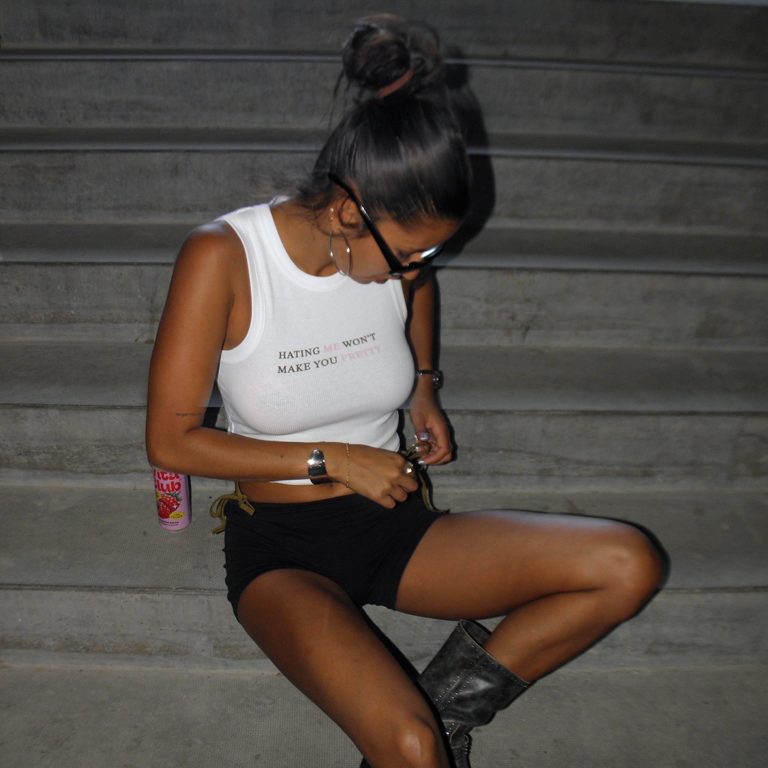 Woman sitting on stairs wearing a white tank top and black shorts, looking at her phone.